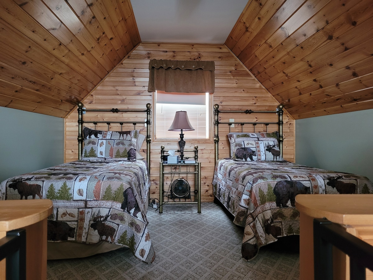 A loft bedroom features two beds with patterned comforters depicting wildlife and nature scenes. A window allows natural light to fill the space, complemented by soft wall and ceiling wood paneling. A bedside table with a lamp is positioned between the beds.