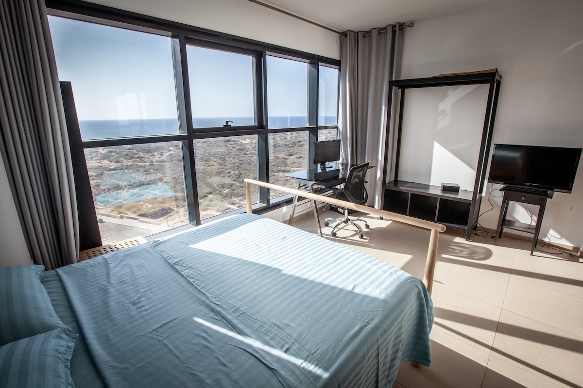 A spacious bedroom is highlighted by large windows offering panoramic sea views. A queen-sized bed with light blue bedding, a desk with a chair, and a TV are present. The room has a minimalist design with natural light flooding in, complemented by neutral-toned walls and flooring.