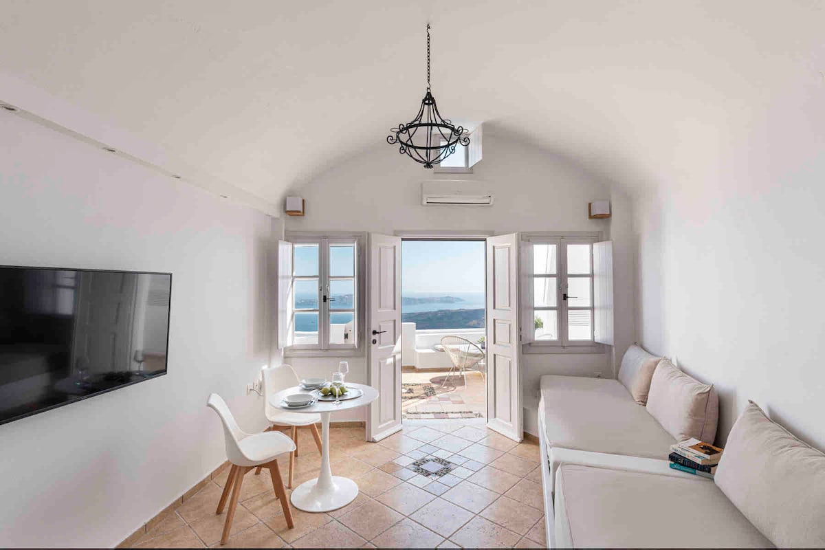 A light-filled living area is presented with a comfortable white sofa and a round dining table. Large doors open to a private terrace, revealing expansive sea views. A wall-mounted TV and a chandelier enhance the inviting ambiance in this elegantly designed space.