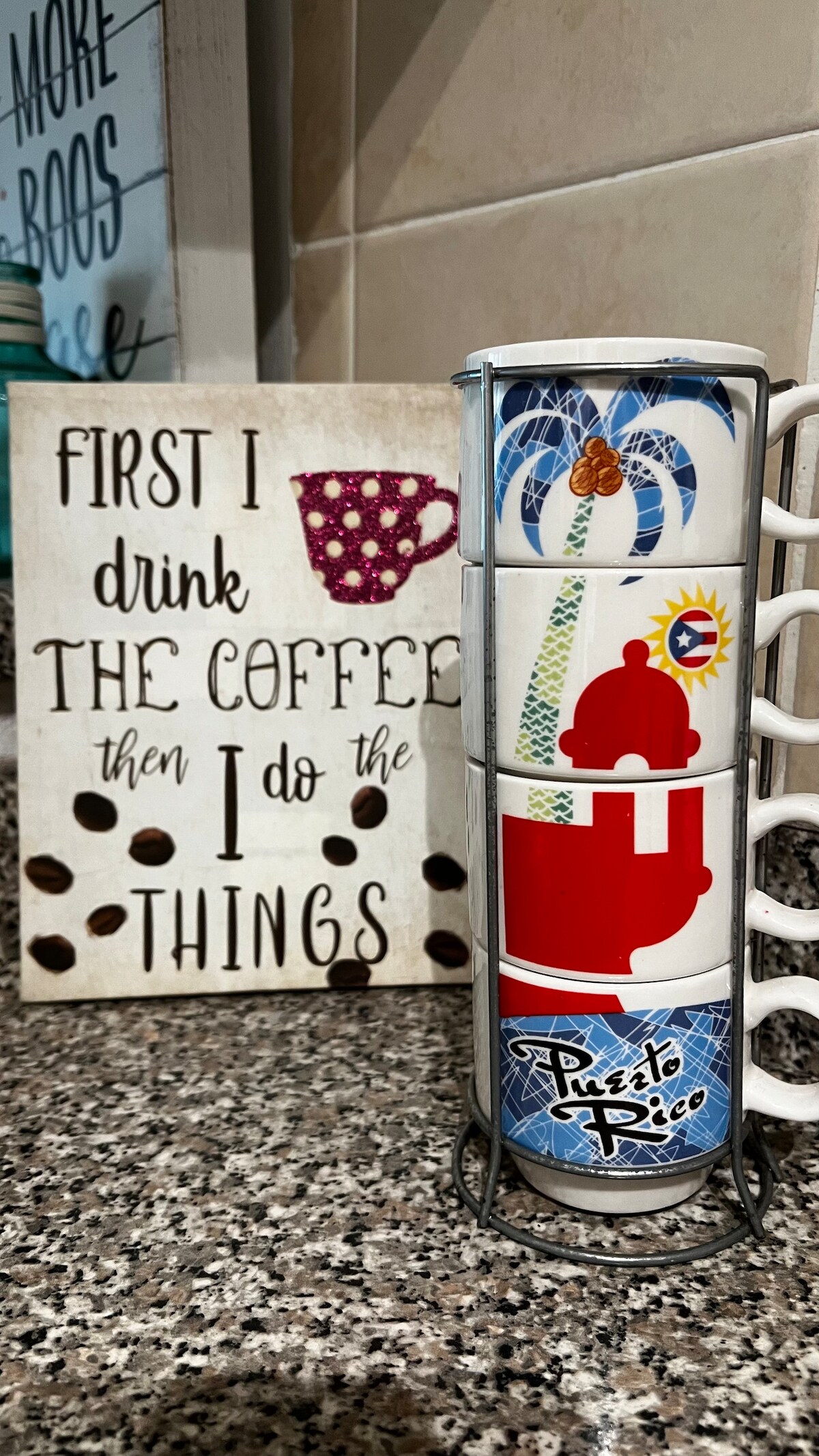 A stack of four decorative coffee mugs is displayed in a metal holder, each showcasing unique designs representing Puerto Rico. A sign with text is positioned nearby, emphasizing the importance of coffee in a lighthearted manner, set against a granite countertop.