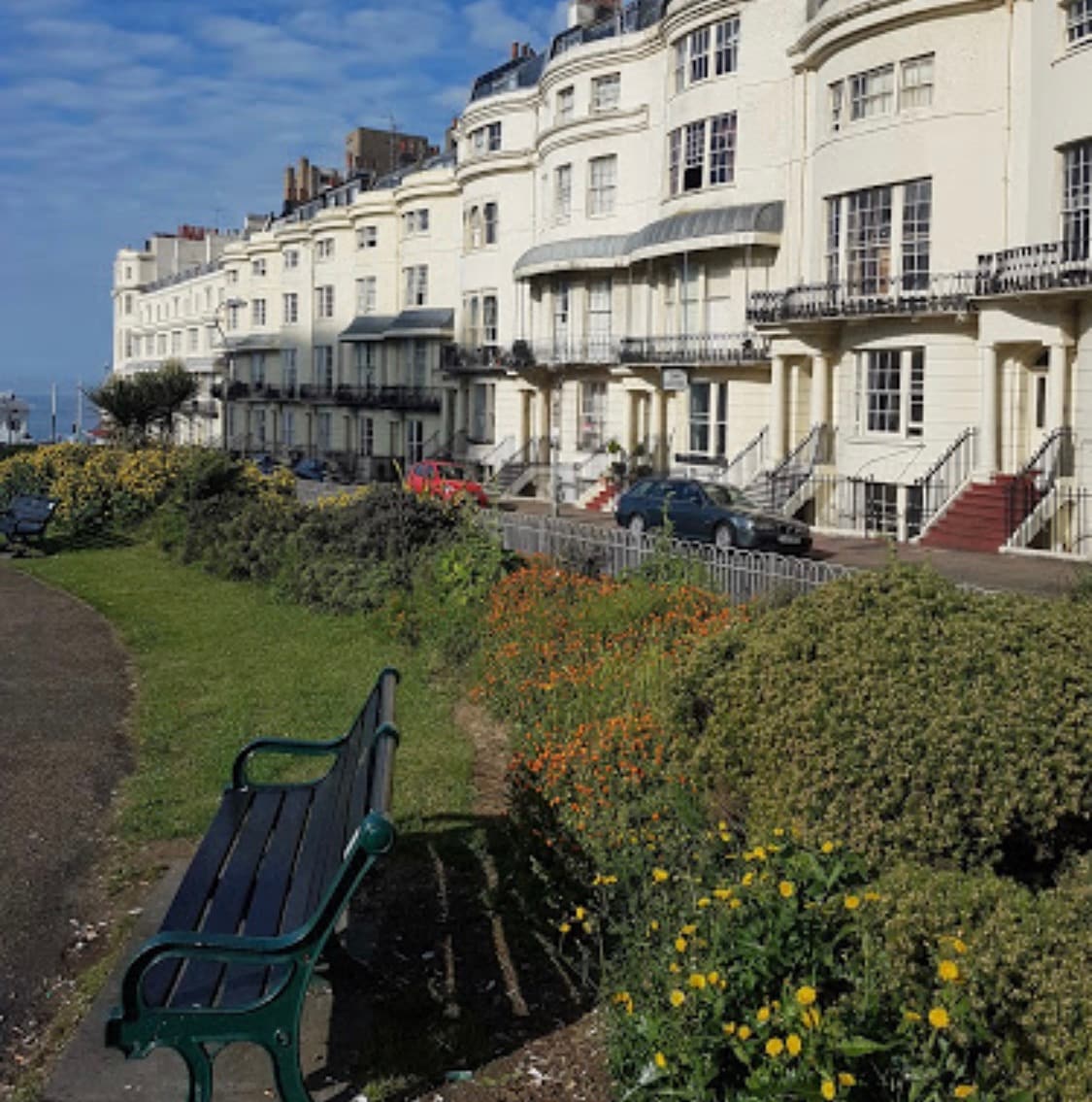 A row of elegant white buildings lines a scenic seafront walkway, featuring balconies and large windows. Colorful flowers and greenery create a pleasant landscape in the foreground, with a solitary bench offering a place to sit and enjoy the view of the sea.