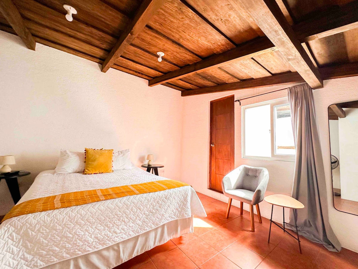 A well-lit bedroom features a spacious bed with a white quilt and a mustard yellow accent pillow. A comfortable armchair is positioned beside a small table, while a window allows natural light to illuminate the room. Exposed wooden beams add warmth to the ceiling.