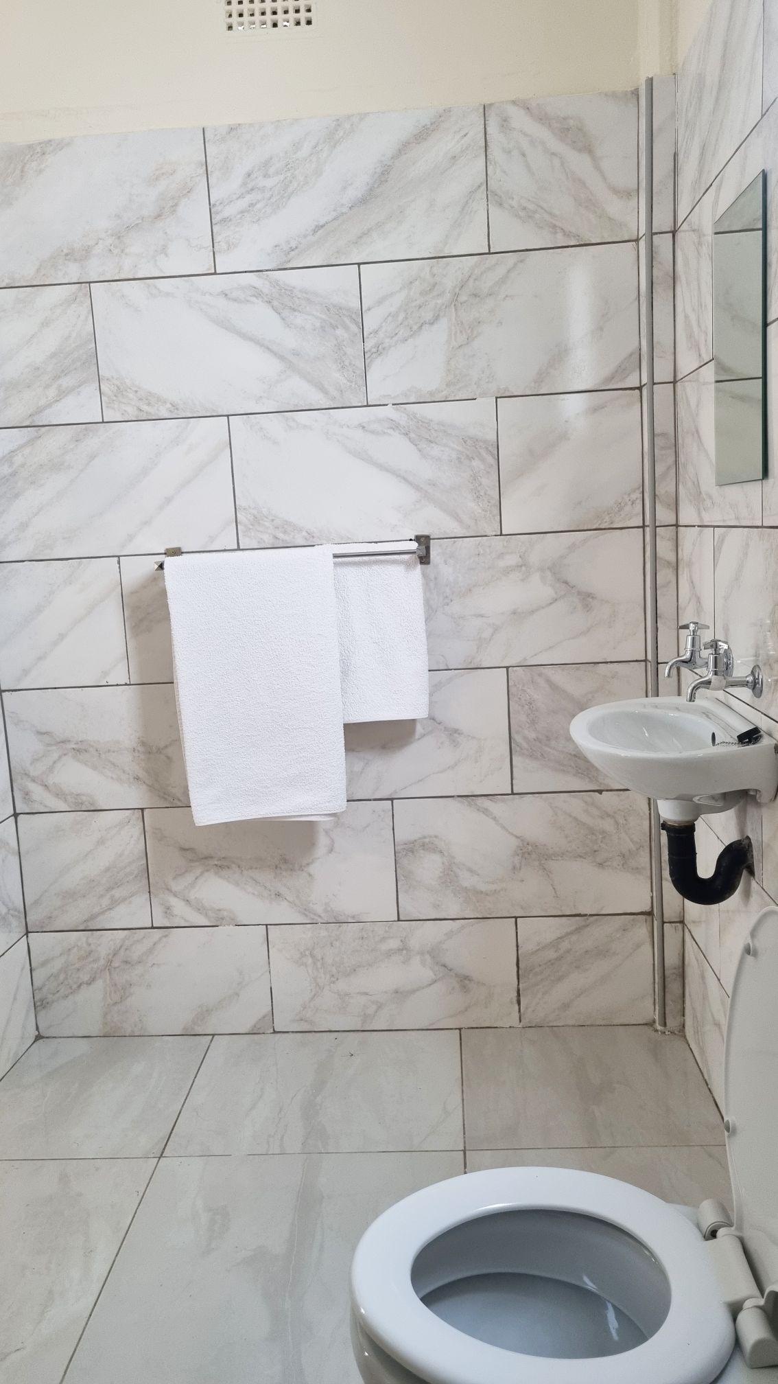 A clean bathroom space features light marble-patterned tiles covering the walls and floor. A white towel hangs on a silver rack, while a small mirror reflects the interior. A modern sink and a toilet are positioned within the room.