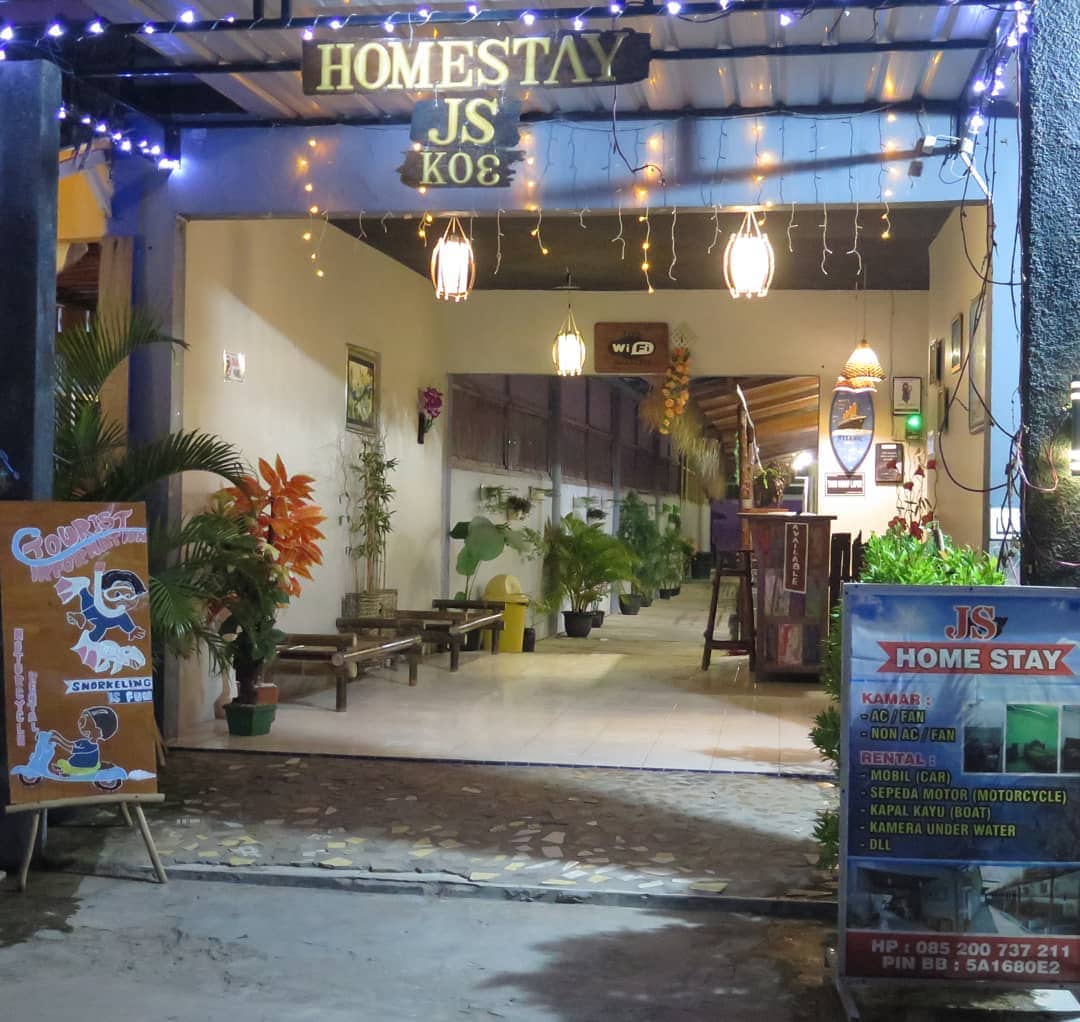 The entrance of Homestay JSKU is framed by warm lighting and plants, creating an inviting atmosphere. A signboard displaying services is positioned on the left, with a clear view of the corridor leading to guest accommodations.