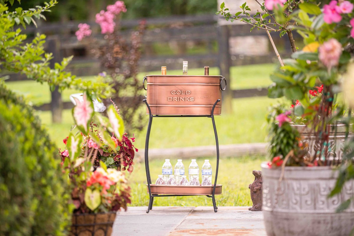 A copper-colored drink cooler is placed on a decorative stand, labeled 'COLD DRINKS.' It sits amidst vibrant flowers and greenery, with water bottles arranged at its base. A background of lush lawns and wooden fencing enhances the outdoor setting.
