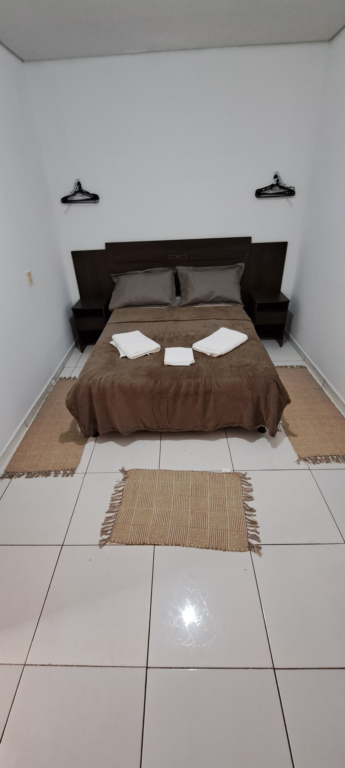 A compact bedroom features a spacious bed with neutral bedding and neatly arranged towels atop it. Two bedside tables stand on either side, with wall hooks positioned above. Light-colored tiles cover the floor, and a small rug adds texture to the room.