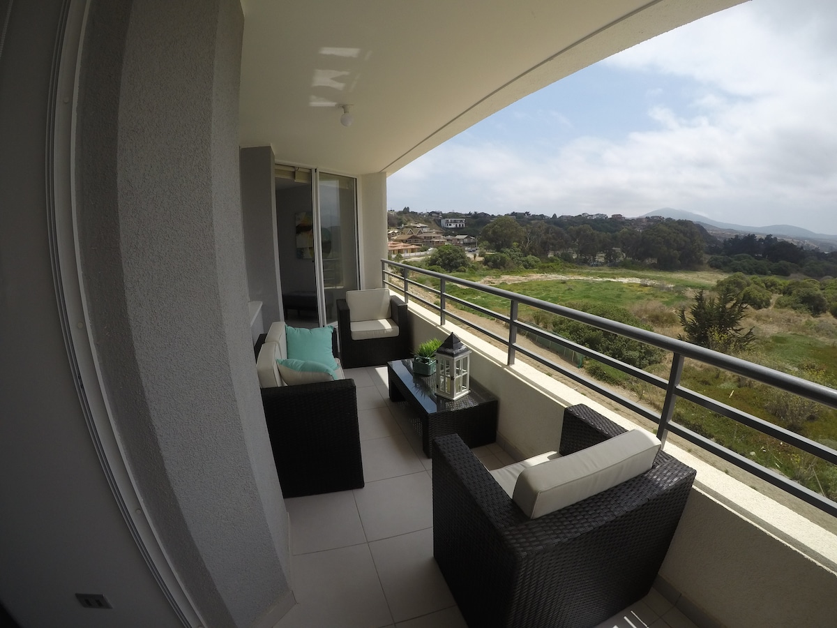 A balcony with modern, dark wicker furniture includes two chairs and a small table, offering a view of the lush surroundings. Large windows allow natural light to fill the space, with soft cushion accents enhancing comfort.