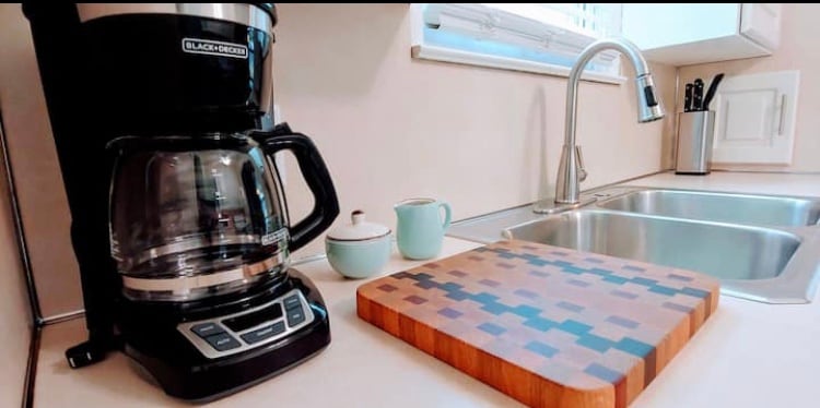 A compact kitchen setup features a black coffee maker alongside a wooden cutting board. Two light blue mugs and a small container are positioned next to the sink, which includes two basins. Natural light enters through a nearby window, brightening the clean counter space.