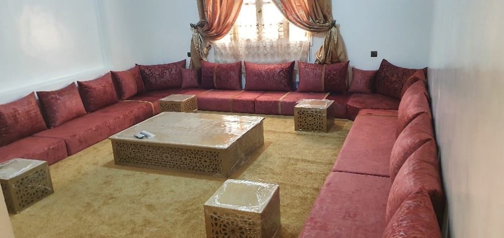 A spacious seating area features a large U-shaped sofa adorned with plush reddish cushions. A low, intricately designed coffee table sits at the center, surrounded by smaller tables. Warm drapes frame the window, allowing soft light to enhance the inviting atmosphere.
