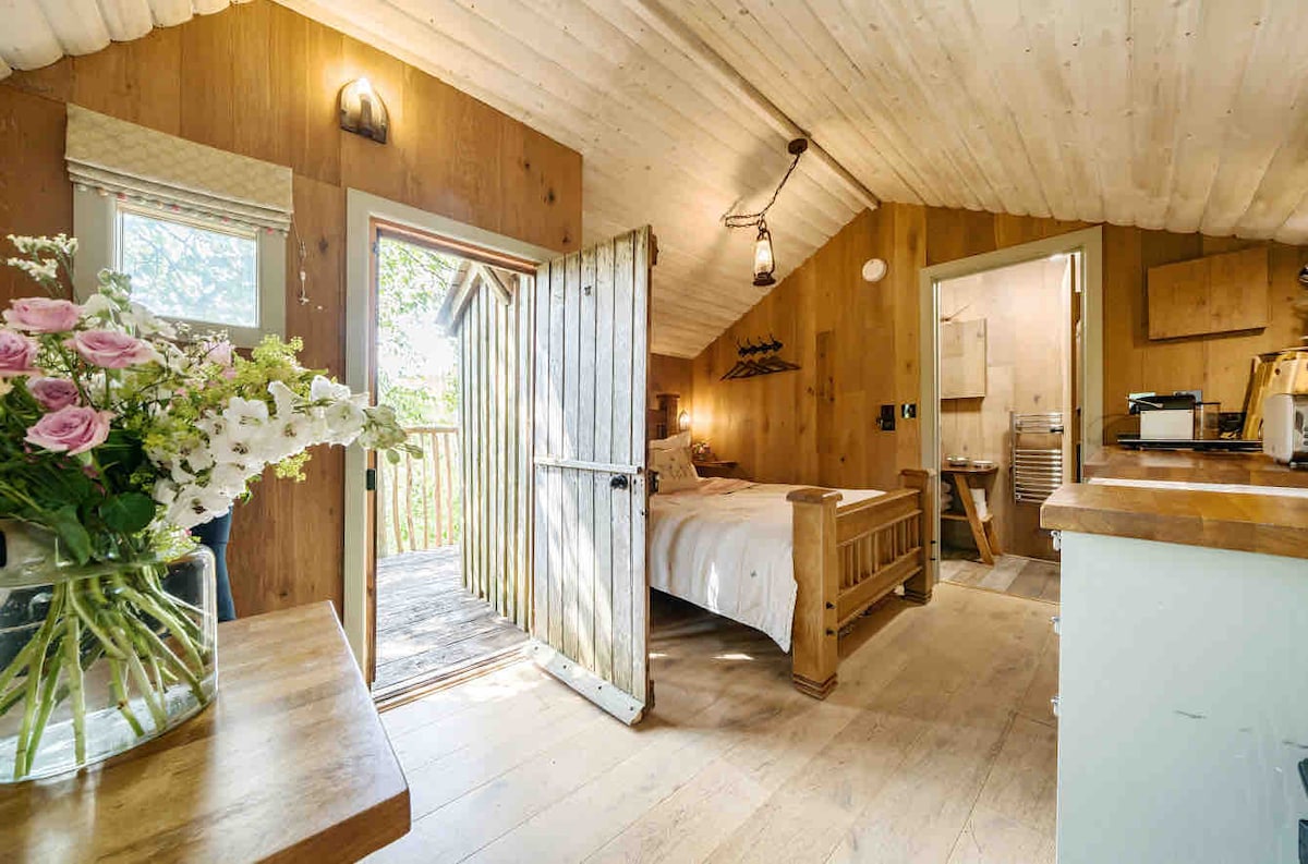 The interior of a rustic treehouse features warm wooden walls and a sloped ceiling. A solid oak bed stands invitingly near an open double door that opens to a deck. A flower vase on a wooden table adds a touch of nature, complemented by natural light from the windows.