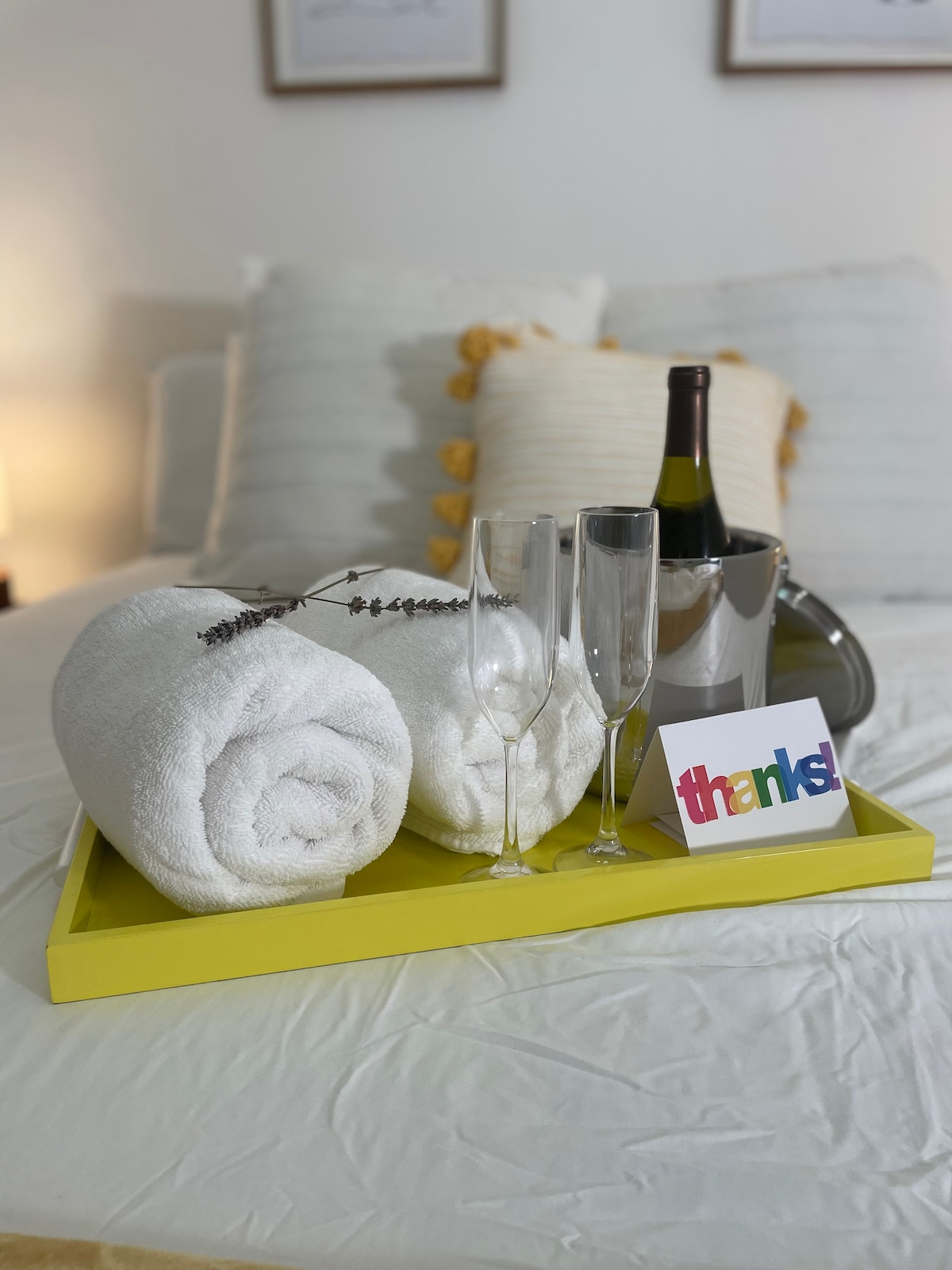 A tray is arranged on a bed featuring rolled white towels, two champagne flutes, and a wine bottle in an ice bucket. A colorful card with the word 'thanks!' is placed alongside the glasses, creating a welcoming touch.