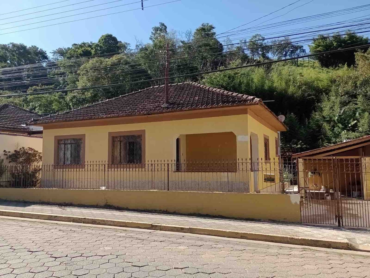 The yellow house is showcased from the street, featuring a tiled roof and multiple windows. A wrought iron fence encloses the front yard, with greenery visible in the background. Nearby, a carport can be seen, providing shelter for vehicles.