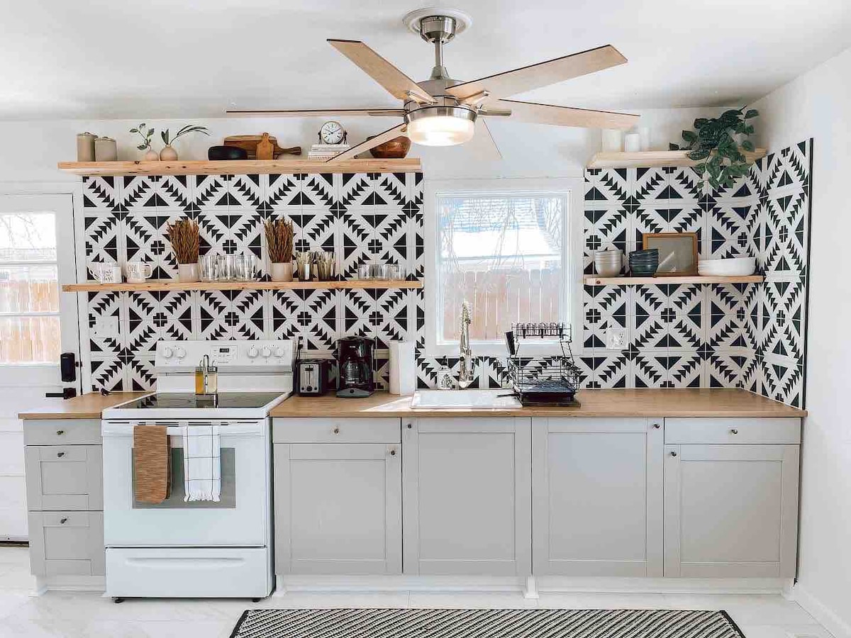 A modern kitchen is presented with a patterned black and white backsplash. Light grey cabinetry and wooden countertops are complemented by stainless steel appliances. Open shelves display kitchen essentials and decorative items. A ceiling fan adds comfort to the space, while a rug enhances the flooring.