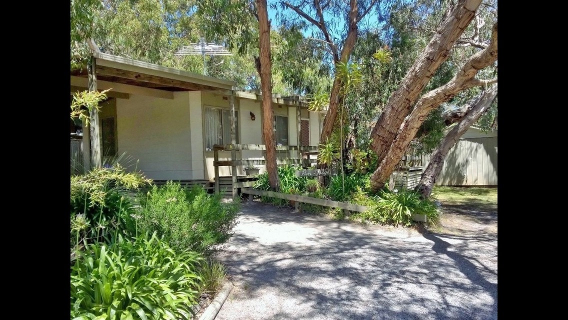 The exterior of the holiday cottage is framed by native trees and lush greenery. The structure features a light-colored facade with a front porch visible, inviting guests to enjoy the natural surroundings. A gravel path leads to the entrance, enhancing the tranquil setting.