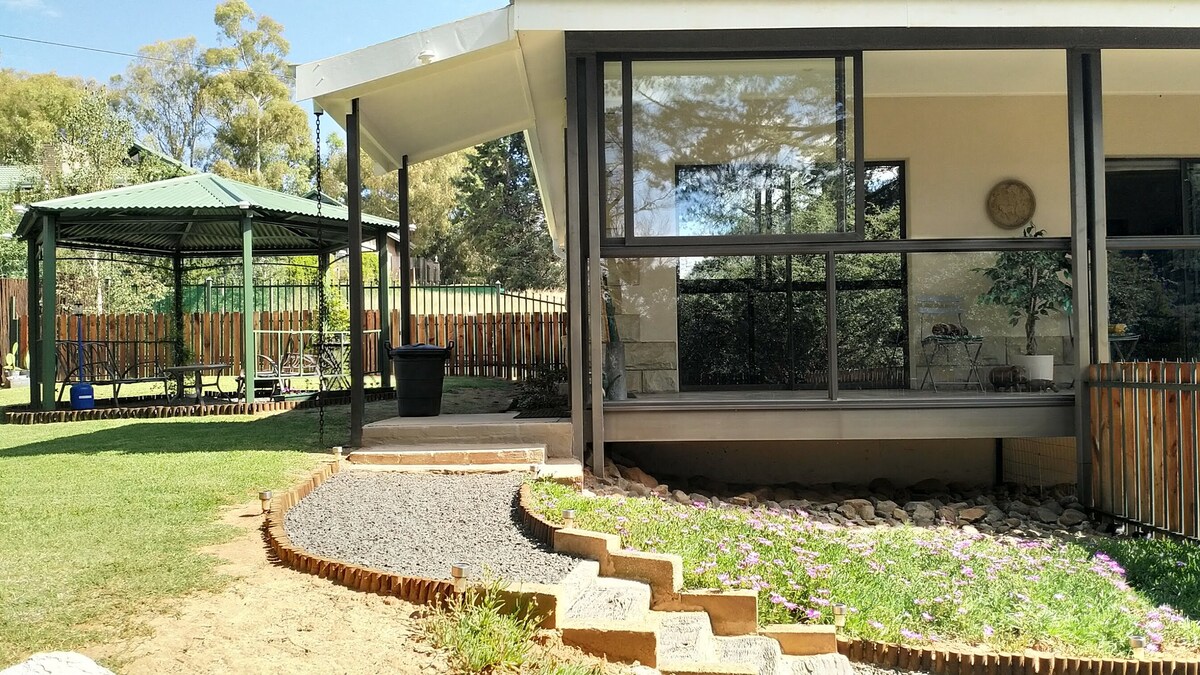A self-catering apartment is pictured, featuring large glass windows that provide natural light. A well-maintained garden with a stone pathway leads to an outdoor gazebo, surrounded by a wooden fence. The grassy area includes scattered small flowers, enhancing the outdoor space.