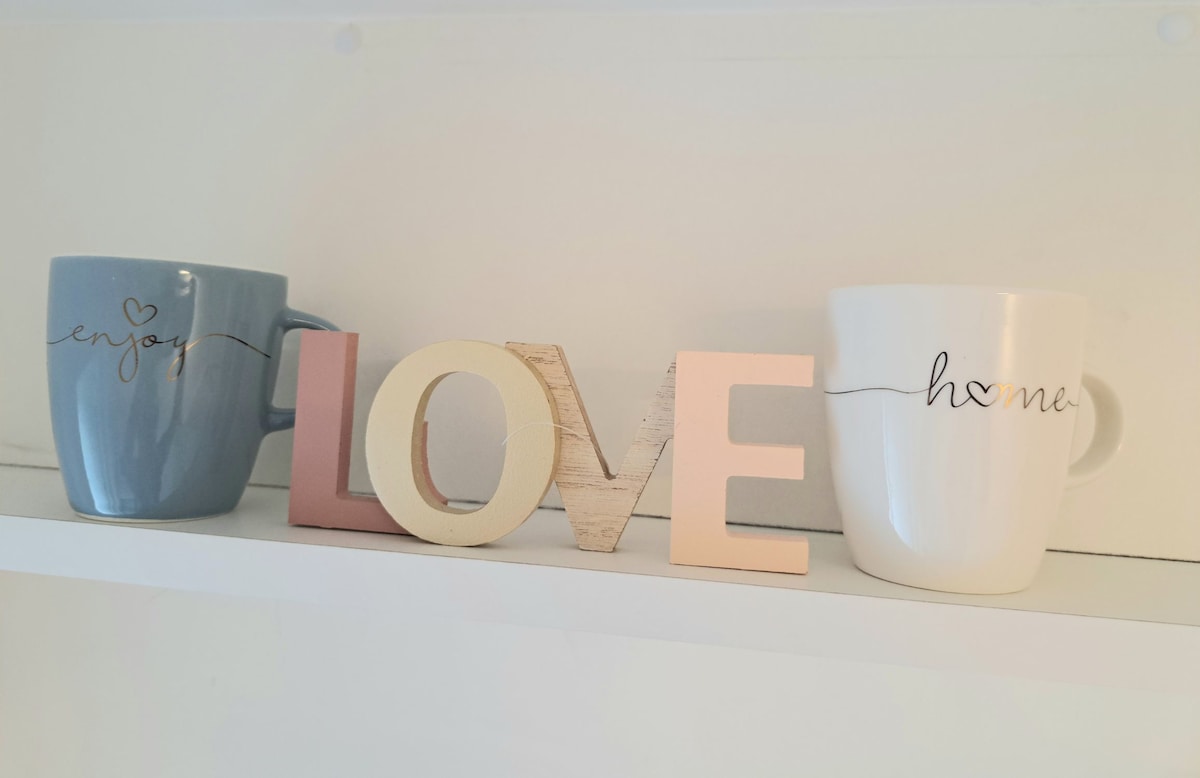 A shelf displays two coffee mugs, one blue and one white, each adorned with a simple phrase. Between them, a light wooden decoration spells 'LOVE' in varying shades, contributing to a cozy and welcoming atmosphere.