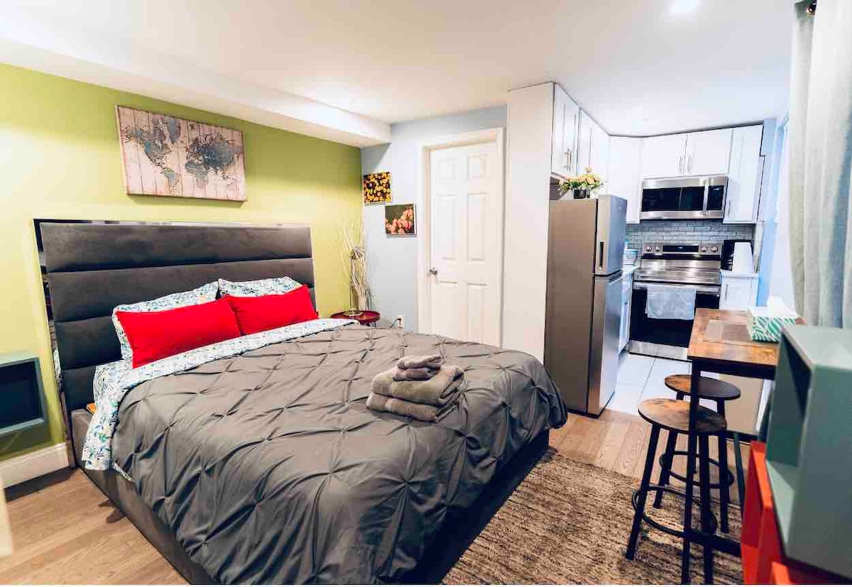 A cozy studio bedroom includes a spacious bed with a gray comforter and decorative pillows. The kitchenette is equipped with modern appliances, and a small dining table with stools provides a comfortable eating area. A subtle green accent wall adds a pop of color.
