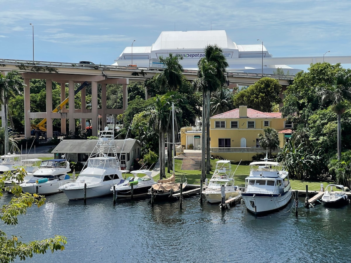 A view of the Miami River reveals several boats docked along the water's edge. Lush greenery surrounds the area, while a light-colored building stands in the background, partially obscured by palm trees. A large white structure is visible above the river.