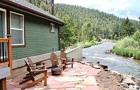 A flagstone patio is positioned alongside a flowing river, featuring several wooden Adirondack chairs arranged for relaxation. Lush green hills are visible in the background, framed by trees, creating a serene outdoor setting.