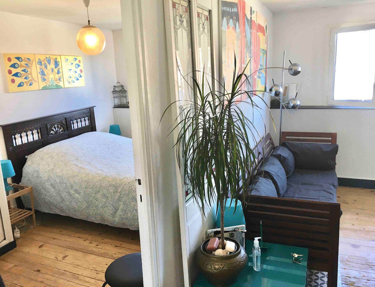 A spacious bedroom features a large bed with a textured blanket, complemented by colorful artwork on the walls. A cozy seating area with a dark sofa is included, alongside a decorative plant. Natural light is provided through a small window, enhancing the open feel of the space.