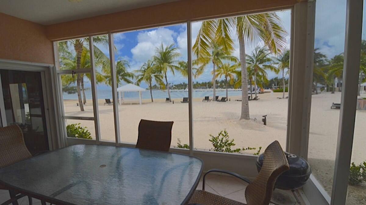 A screened lanai features a glass-top table and comfortable chairs, offering a view of the beach and palm trees outside. The sandy shoreline leads to calm waters, with lounge chairs arranged under shaded canopies in the background, creating a serene coastal atmosphere.