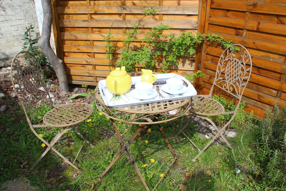 A quaint outdoor seating area is featured, with two intricately designed metal chairs positioned around a small, round table. A yellow teapot and cups are arranged on a white tray, set against a backdrop of wooden fencing and greenery.