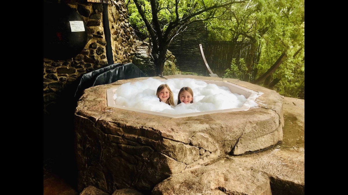 A hot tub is nestled within a stone enclosure, surrounded by leafy trees and natural greenery. Plentiful bubbles create a calming atmosphere as they rise above the water's surface, inviting guests to relax in this outdoor space.