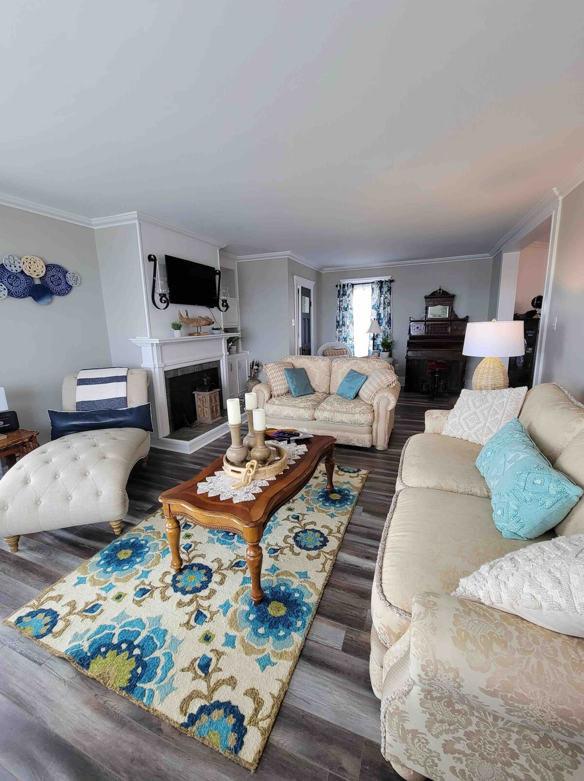 A bright and inviting living room features a stylish beige sofa and a coordinating chaise lounge, arranged around a wooden coffee table adorned with decorative candles. A vibrant area rug with floral patterns adds color, while a flat-screen TV hangs above a classic fireplace.