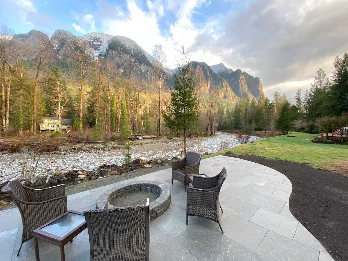 A stone patio features four wicker chairs arranged around a circular fire pit, facing the gently flowing Snoqualmie River. Lush greenery and tall trees border the riverbanks, while towering mountains rise in the background under a softly lit sky.