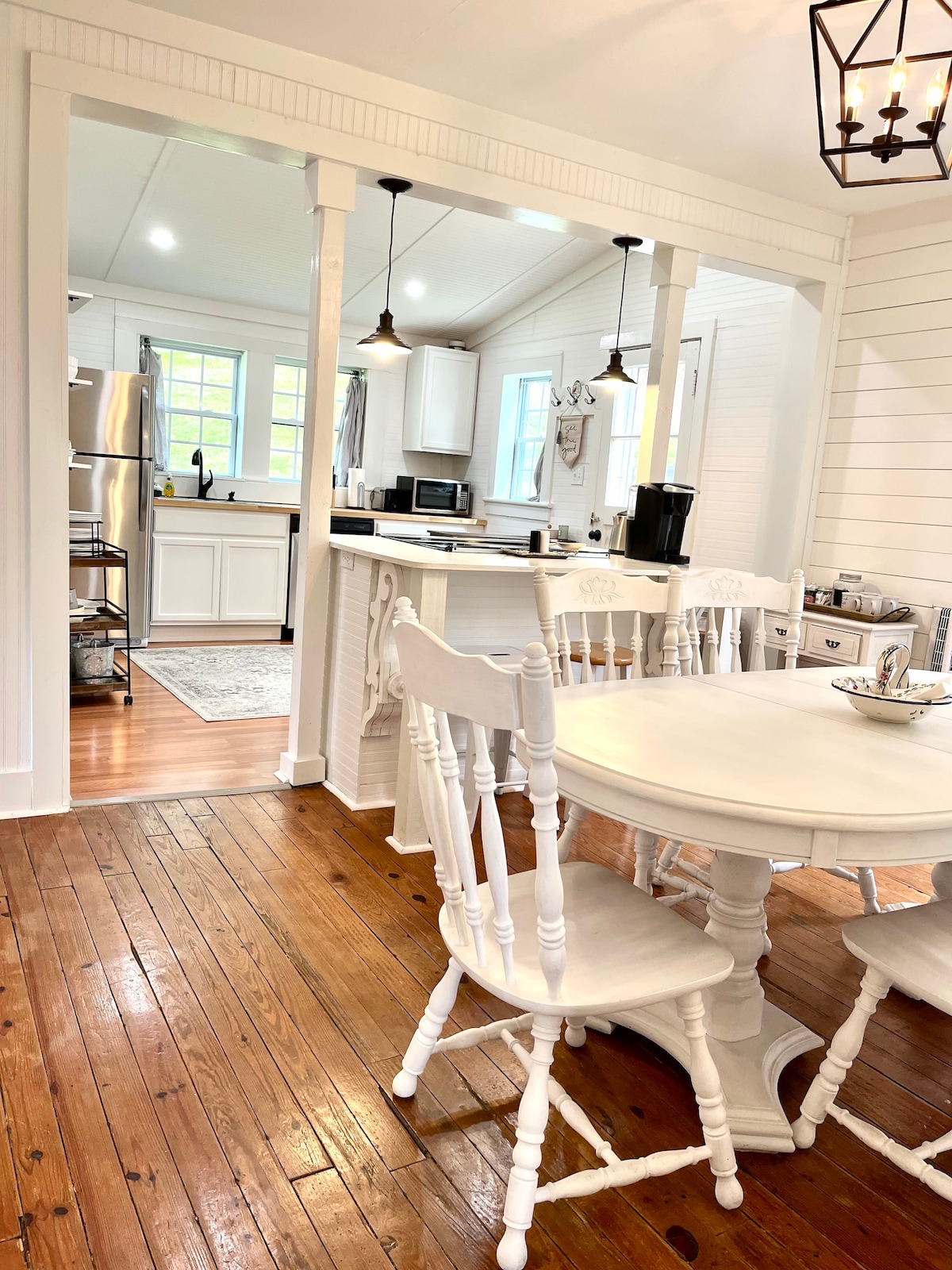 The kitchen area features a light wood dining table surrounded by four white chairs. A spacious counter with modern appliances is visible in the background. Natural light fills the room through multiple windows, highlighting the rustic wooden floors and clean white walls.