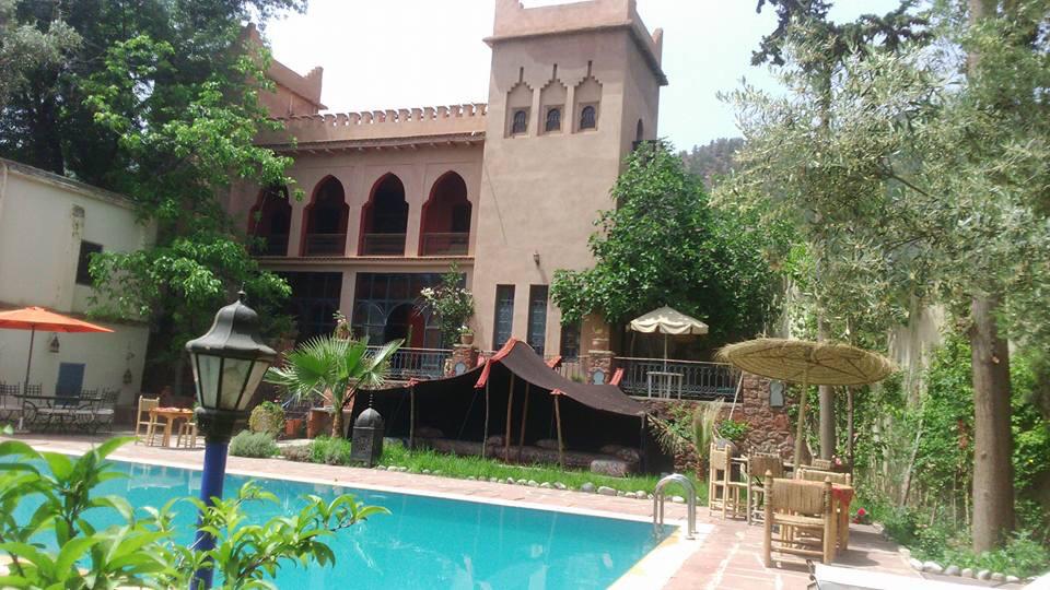 A striking building with traditional Moroccan architecture is surrounded by greenery. A clear pool reflects the sun, while shaded seating areas invite relaxation. A tent structure and patio umbrellas provide a comfortable outdoor space, enhancing the serene atmosphere of the garden.