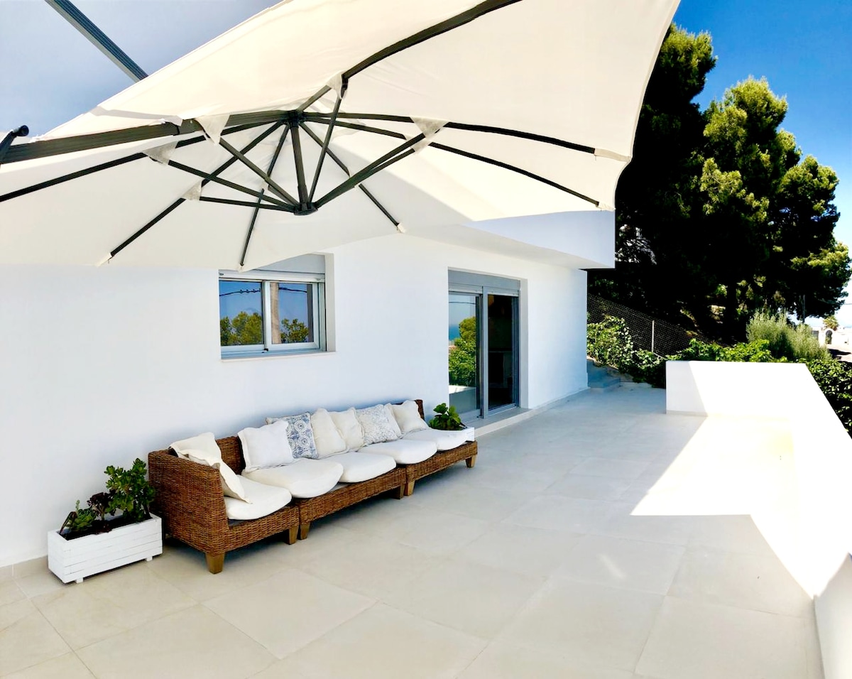 A spacious terrace features a large umbrella providing shade over a comfortable sofa with white cushions. The area is set against a backdrop of greenery and a modern white wall, creating an inviting space for relaxation.