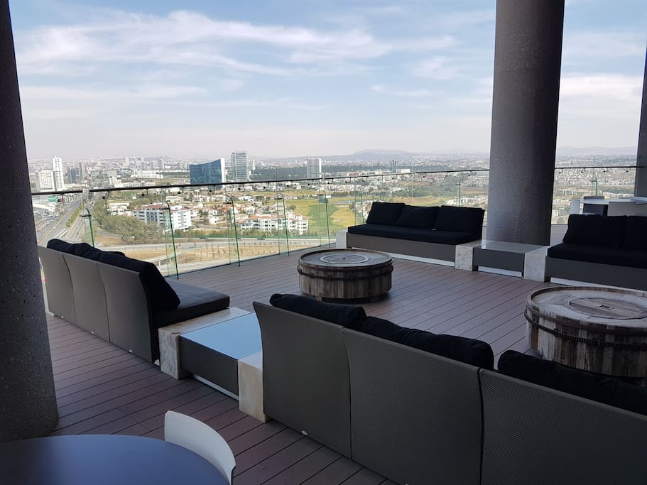 A spacious terrace area features comfortable seating arrangements, including several cushioned sofas and occasional tables. The view showcases the surrounding cityscape and greenery, with expansive glass railings offering unobstructed sightlines. The flooring is wooden, enhancing the open and inviting atmosphere.