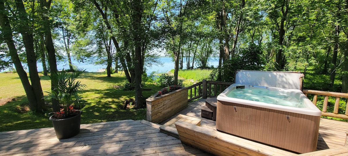 A spacious outdoor deck features a hot tub surrounded by greenery. In the background, the tranquil view of Mississippi Lake is visible through the trees, enhancing the serene atmosphere. Potted plants add a touch of nature to the deck space.
