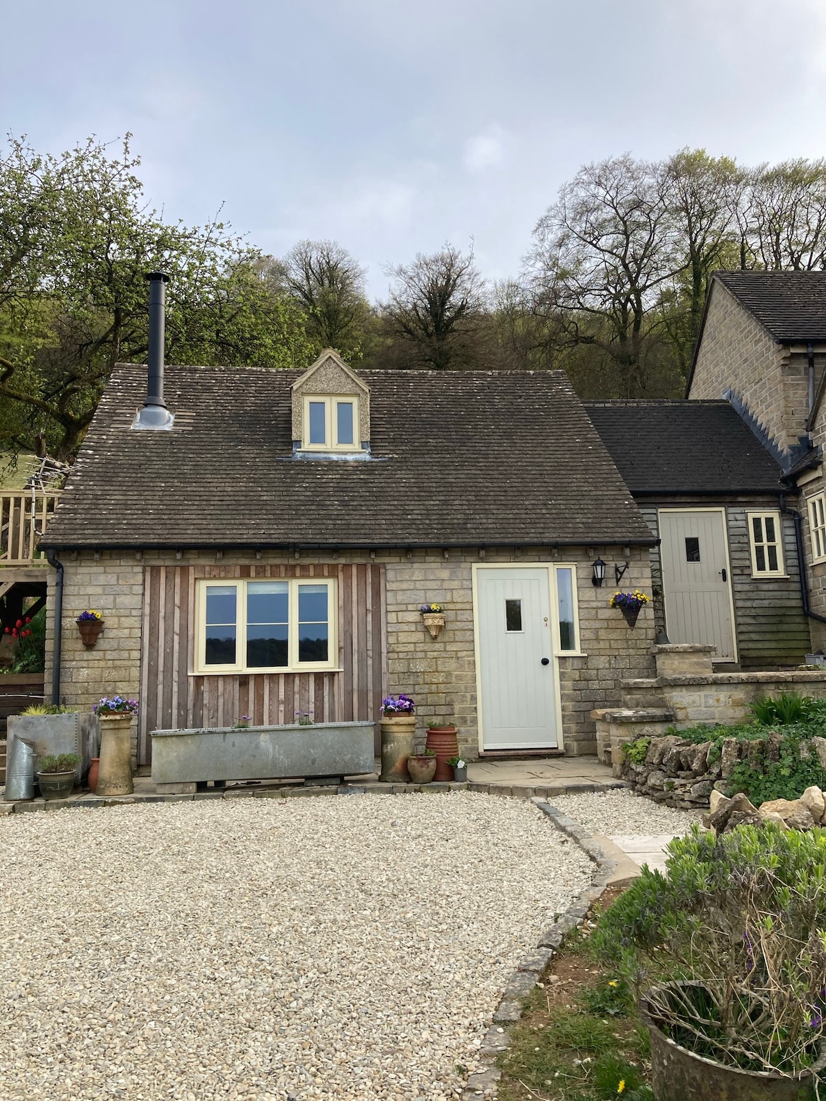 The exterior of a charming stone annexe is shown, featuring a pitched roof and a white door. Planters with colorful flowers add a welcoming touch, while surrounding gravel pathways and lush greenery create a serene rural setting.