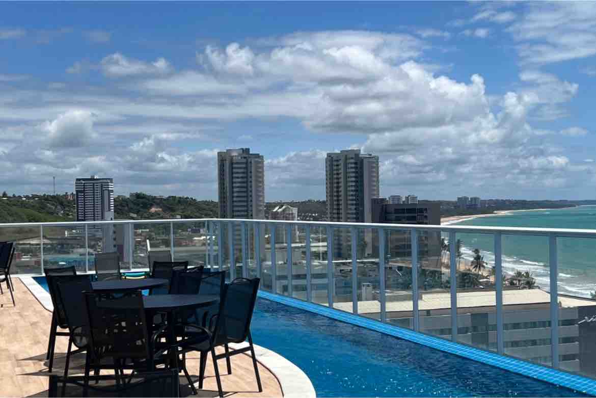 An outdoor pool area is visible, featuring several black tables and chairs arranged near the edge. The scene includes a panoramic view of the beach and tall buildings in the distance, with a clear blue sky and soft clouds overhead.