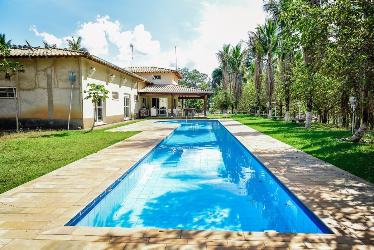 A long swimming pool is surrounded by tiled pathways and lush greenery. The pool reflects the clear sky above, while a spacious outdoor seating area is visible in the background beneath a shaded porch. Palm trees add to the serene atmosphere.