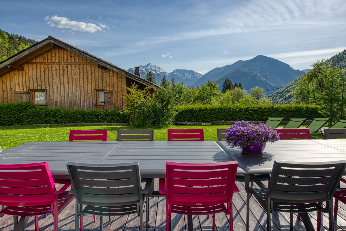A large outdoor terrace features a long dining table surrounded by colorful chairs, providing ample seating. Lush green grass and blooming flowers enhance the area. In the background, mountains and a wooden chalet can be seen, creating a serene setting for outdoor meals.
