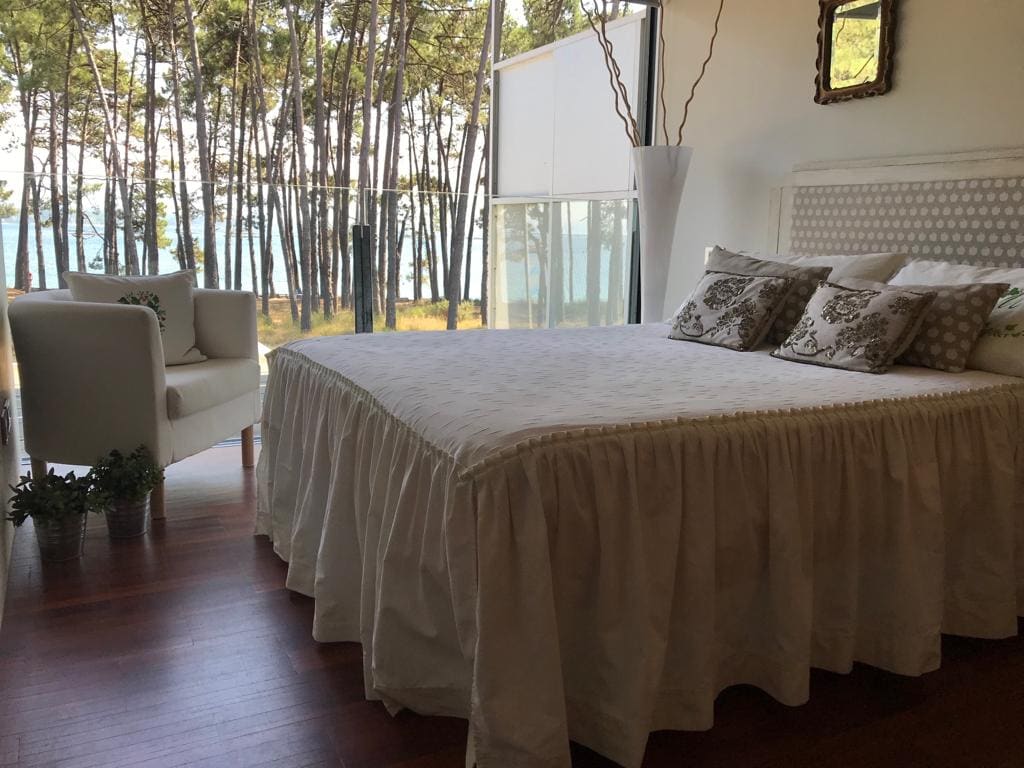 A bright bedroom features a spacious bed with a ruffled coverlet, complemented by decorative cushions. A cozy armchair is positioned nearby, offering views of tall trees and a glimmering body of water through large glass windows, inviting natural light into the space.