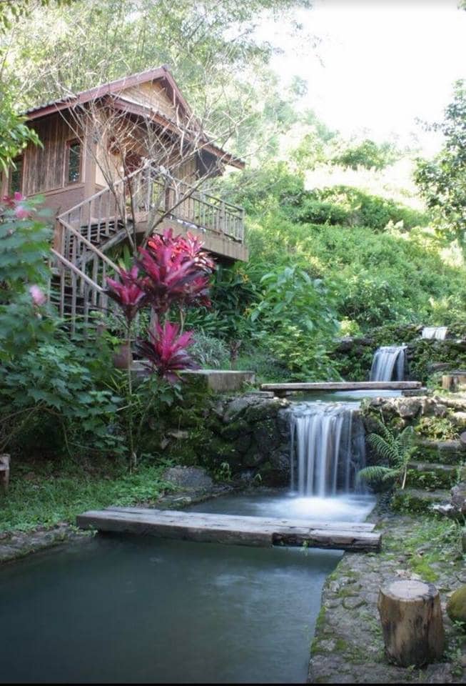 A wooden house is nestled among lush greenery, accompanied by cascading waterfalls and vibrant tropical plants. A tranquil pond reflects the natural surroundings, with a wooden walkway providing access. The setting evokes a serene and private atmosphere, ideal for relaxation.