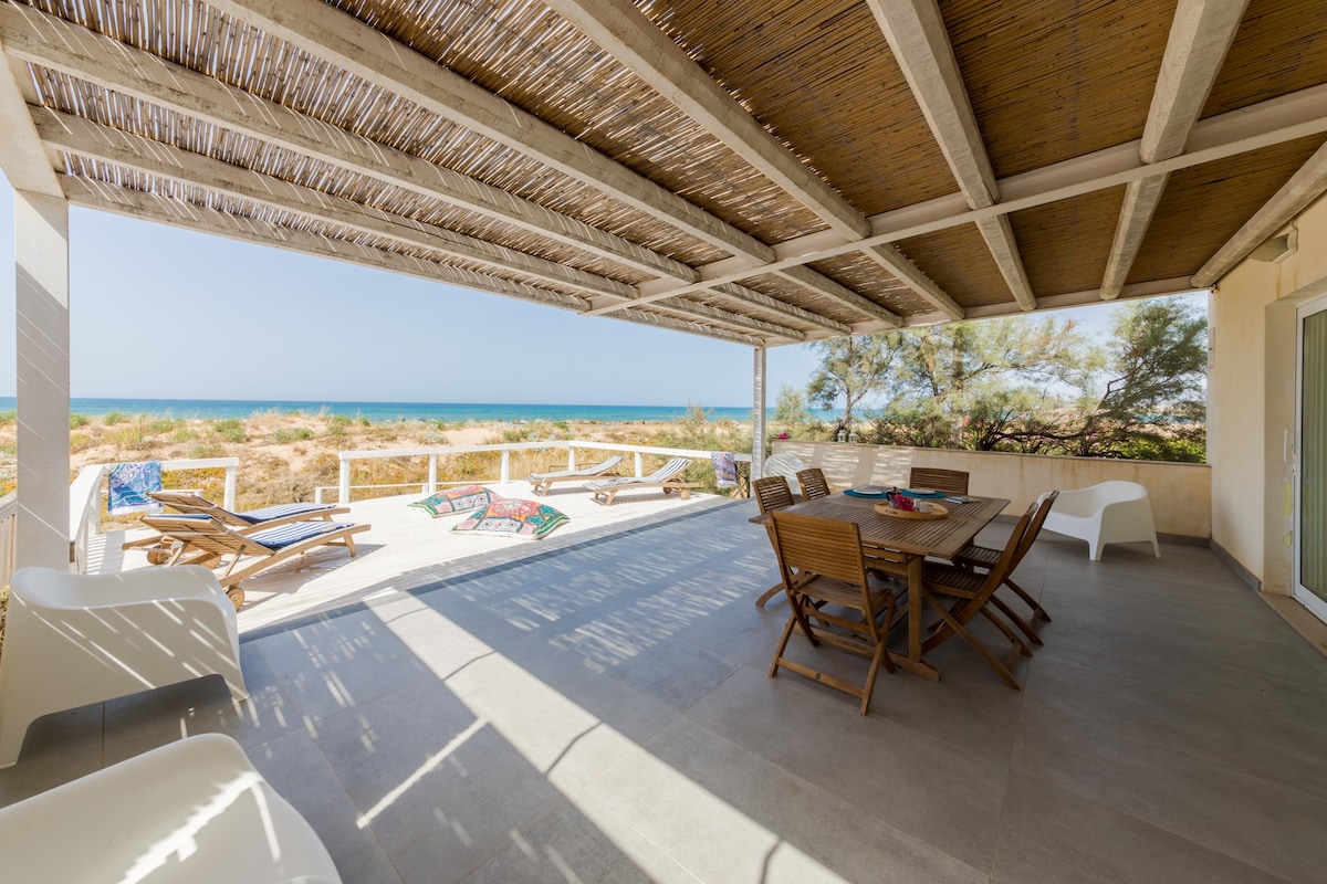 A covered outdoor terrace provides shade and features a wooden dining table surrounded by chairs. Sun loungers are visible nearby, with a view of the sandy beach and sea in the background. Natural light illuminates the space, creating a relaxed atmosphere.