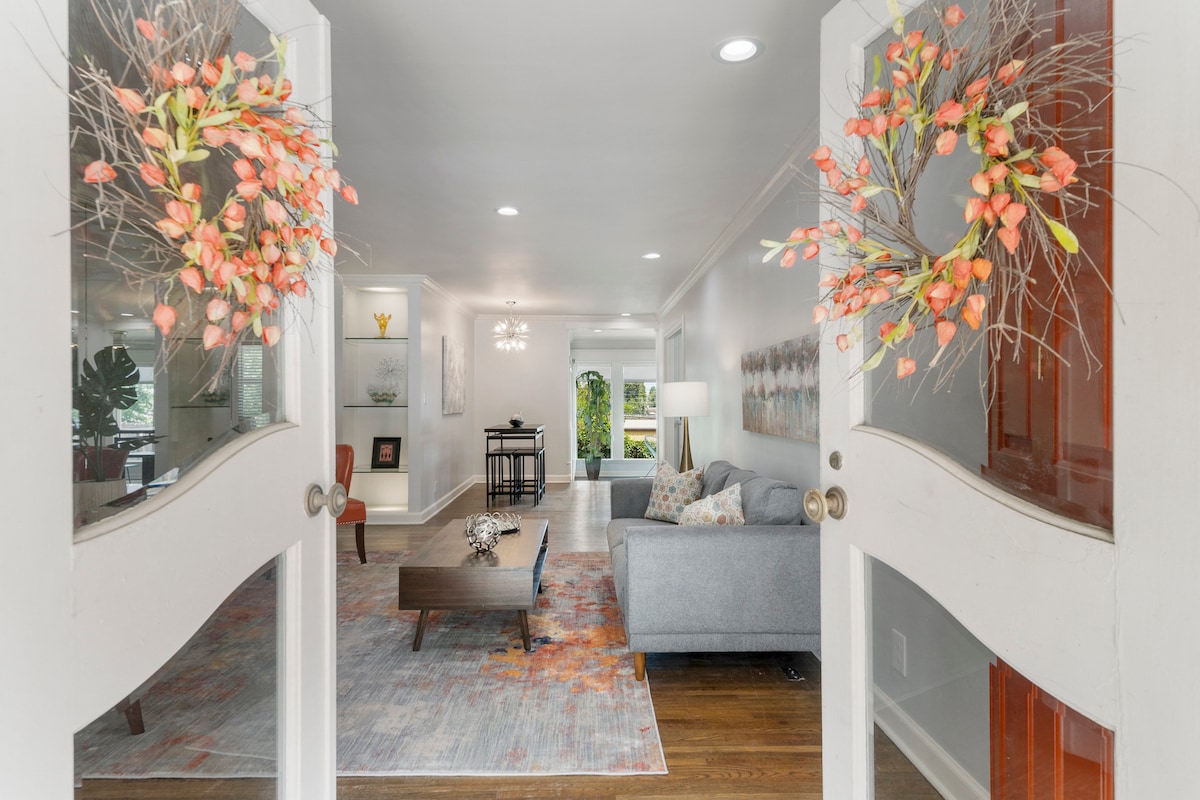 The entrance is framed by double doors adorned with floral wreaths, leading into a bright living area. A gray sofa is positioned opposite a wooden coffee table, complemented by a colorful area rug. Natural light fills the space, enhancing the modern decor.