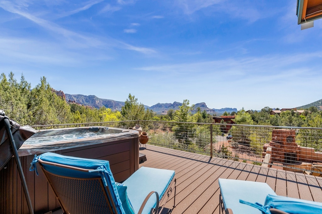 A spacious outdoor deck showcases a hot tub surrounded by serene natural landscapes. Comfortable lounge chairs provide a perfect spot for relaxation, while expansive views of red rock formations and greenery create a tranquil setting under a clear blue sky.