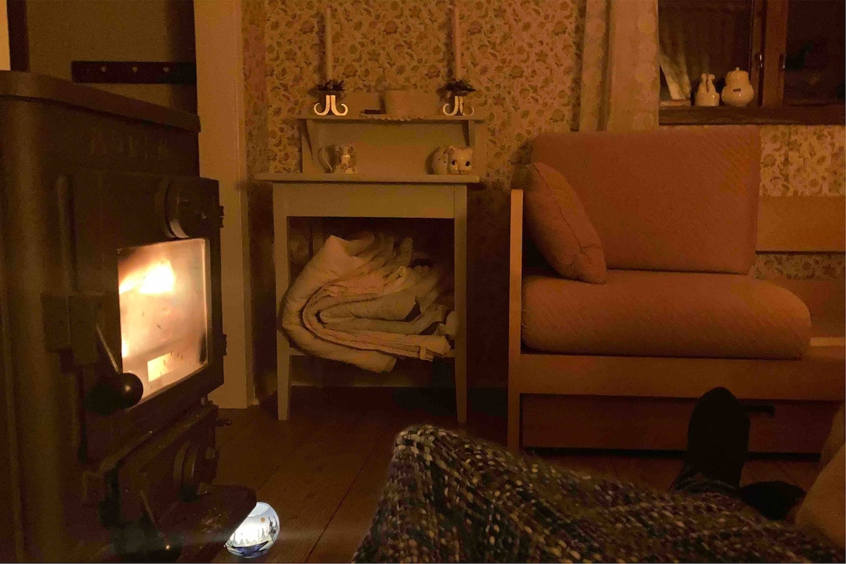 A cozy living space is highlighted by a wood-burning stove, providing warmth and a soothing glow. A soft armchair is positioned near the stove, accompanied by a small side table displaying decorative items. A stack of blankets is neatly stored beneath the table.