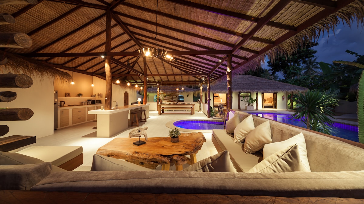The spacious open-plan living area is highlighted by high thatched ceilings and natural wooden beams. A comfortable seating arrangement is positioned around a unique wooden coffee table. A tranquil swimming pool is visible, illuminated softly at night and surrounded by tropical greenery.