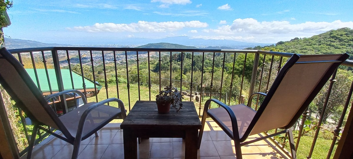 A balcony view showcases two comfortable chairs alongside a wooden table, with a small plant as a centerpiece. The expansive landscape reveals rolling hills and a distant city skyline, highlighted by clear blue skies and scattered clouds.