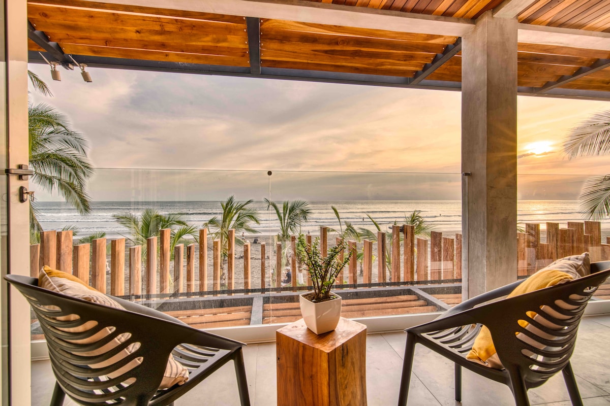 A view from a balcony is presented, featuring two modern chairs set beside a small wooden table. The backdrop showcases the Pacific Ocean and a colorful sunset, framed by swaying palm trees and rustic wooden fencing.