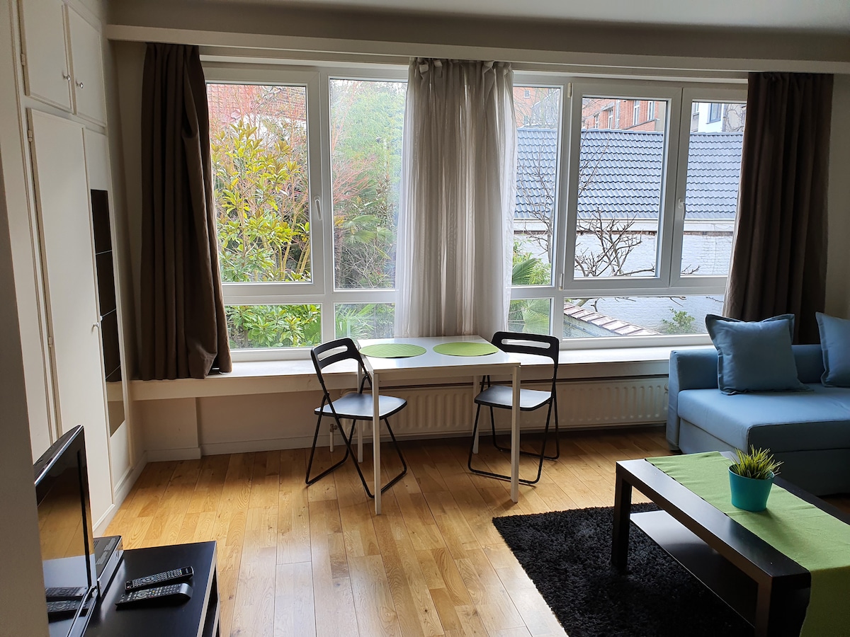 A cozy living area showcases large windows with plant views outside. Two green-accented tables are paired with black chairs, providing a casual dining space. A light blue sofa invites relaxation, positioned next to a coffee table on a dark rug, enhancing the room's warmth.