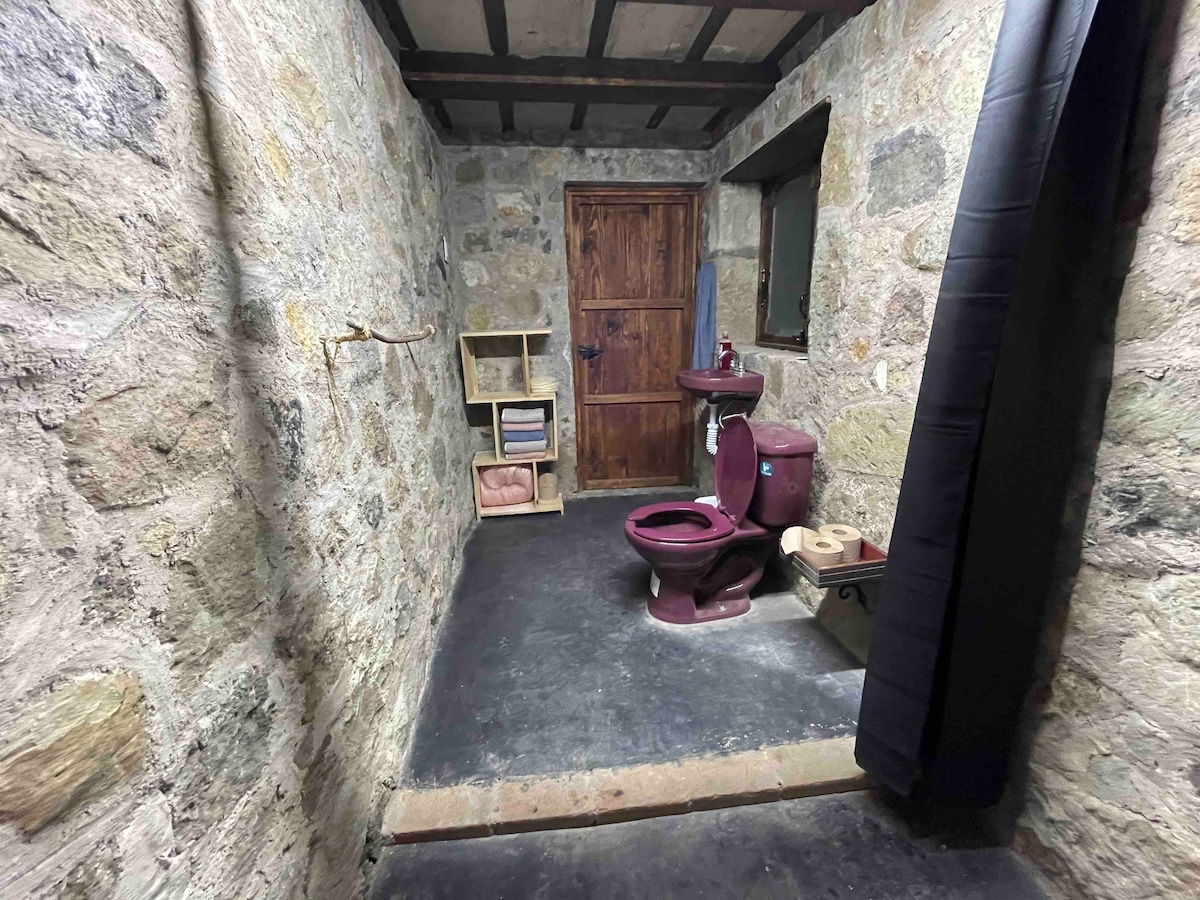 A functional bathroom space is presented, featuring rustic stone walls and a wooden door. A maroon toilet and matching sink are visible, along with small shelving for toiletries. The floor is finished with dark, durable material, contributing to a practical atmosphere.