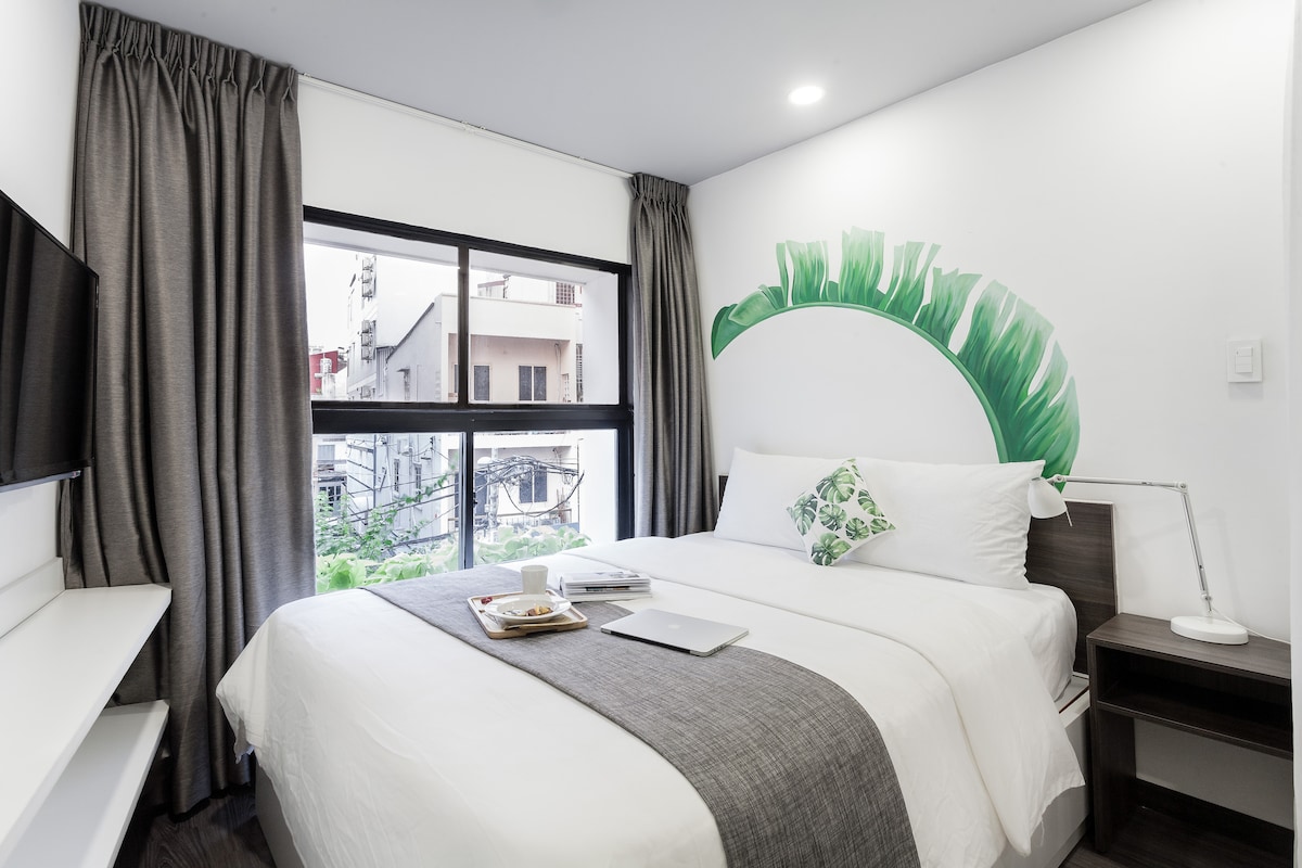 A cozy room features a spacious bed with soft bedding and decorative pillows. A large window is adorned with sheer and heavy curtains, providing natural light. A small desk with a laptop sits nearby, while a minimalist TV hangs on the wall, complemented by a nature-inspired mural.